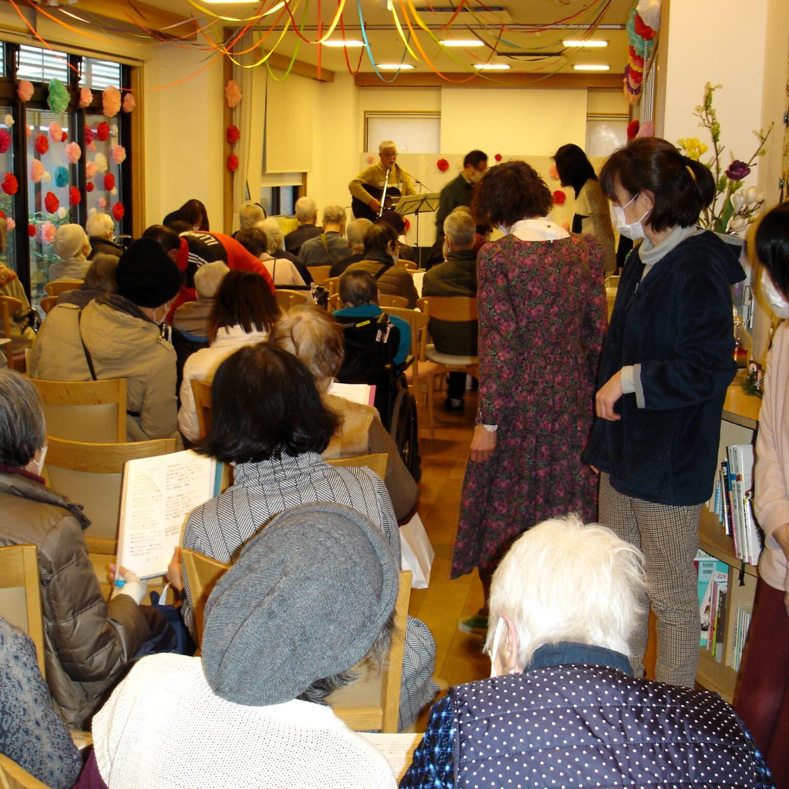 介護施設でのレクリエーション行事。多くの入居者と職員が集まり、音楽演奏を鑑賞している様子。